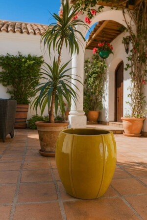 Macetero decorativo de exterior amarillo sobre suelo de barro en patio mediterráneo con plantas.
